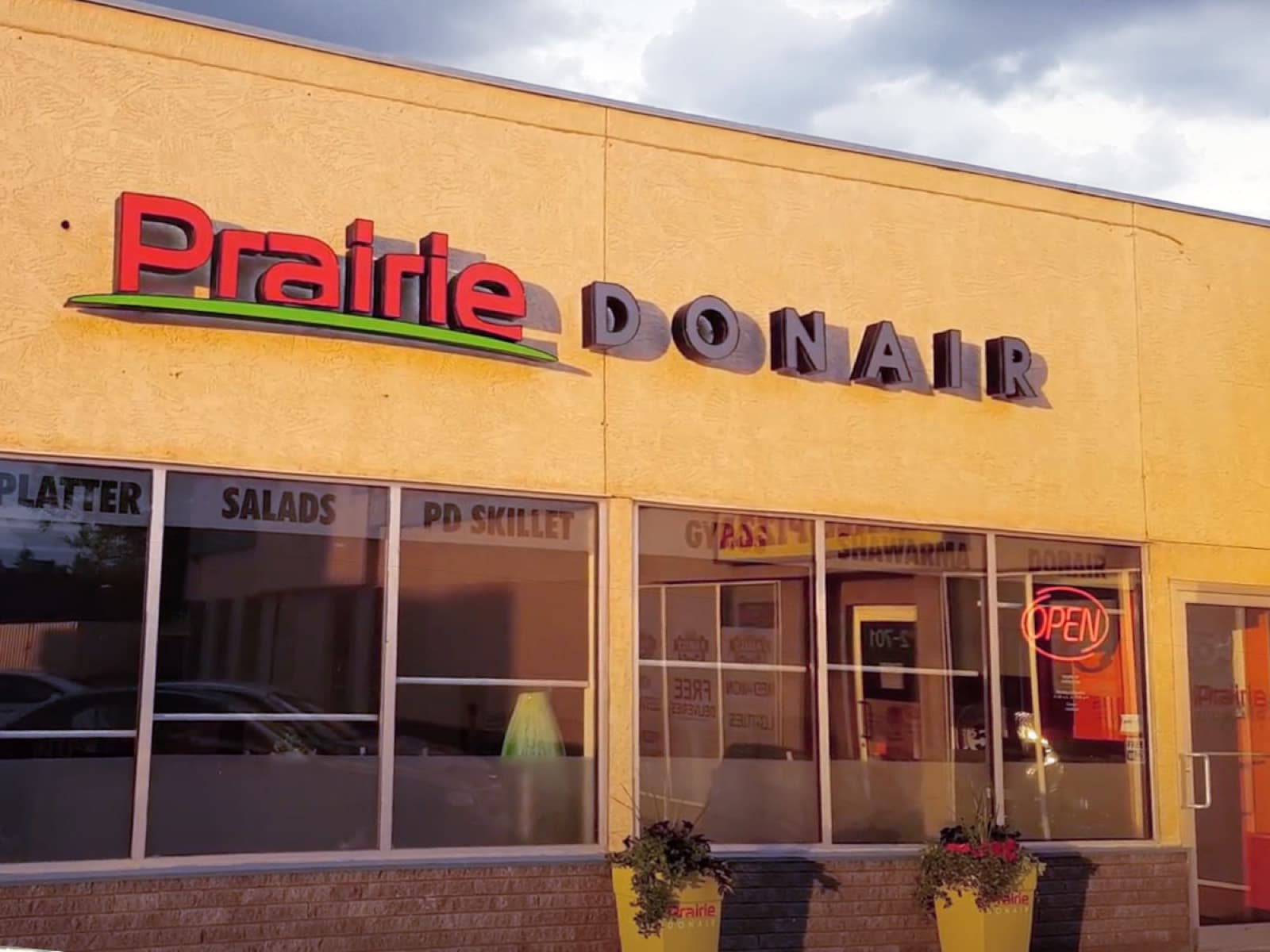 A Prairie Donair storefront on a bright afternoon: the red and lime wordmark mounted above a row of tall windows lettered with the menu — platters, salads, gyros, shawarma — and a lit OPEN sign in the door.